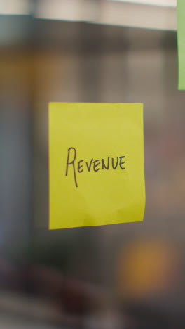 Vertical-Video-Close-Up-Of-Woman-Putting-Sticky-Note-With-Revenue-Written-On-It-Onto-Transparent-Screen-In-Office